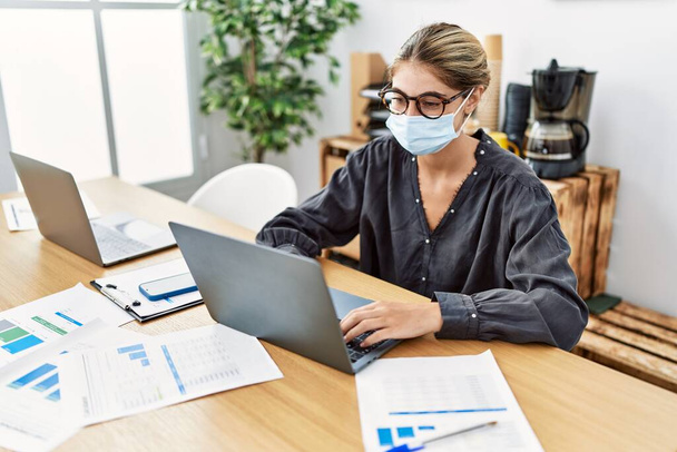 Young blonde woman working at the office wearing safety mask looking positive and happy standing and smiling with a confident smile showing teeth  - Foto, immagini