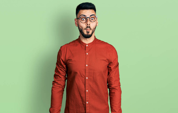 Jonge Spaanse man met baard die een zakelijk hemd draagt en een bril die vissengezicht trekt met lippen, gek en komisch gebaar. grappige uitdrukking.  - Foto, afbeelding