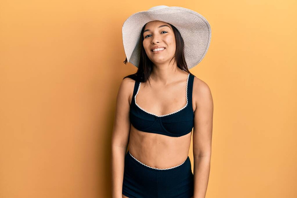 Jeune femme latine portant un bikini et un chapeau d'été regardant positif et heureux debout et souriant avec un sourire confiant montrant des dents  - Photo, image