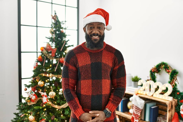 Afrykański Amerykanin w kapeluszu Świętego Mikołaja stojący przy choince ze szczęśliwym i chłodnym uśmiechem na twarzy. szczęśliwy człowiek.  - Zdjęcie, obraz