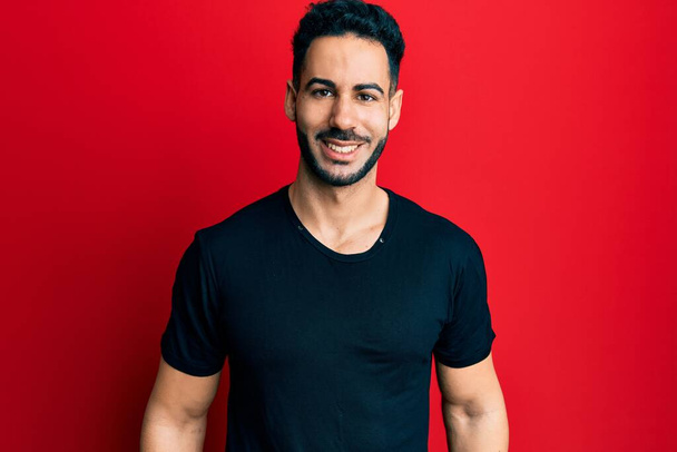 Junger hispanischer Mann in lässigem schwarzem T-Shirt mit einem fröhlichen und kühlen Lächeln im Gesicht. Glück gehabt.  - Foto, Bild