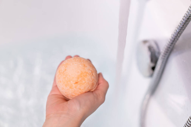 Beautiful woman holding an aromatic orange bath bomb on her hands before put it into the bathtub. Wellness and hydro therapy relaxation concept. - Photo, image