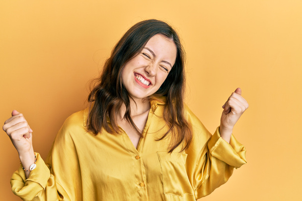 Young brunette woman wearing casual yellow shirt very happy and excited doing winner gesture with arms raised, smiling and screaming for success. celebration concept.  - Photo, Image