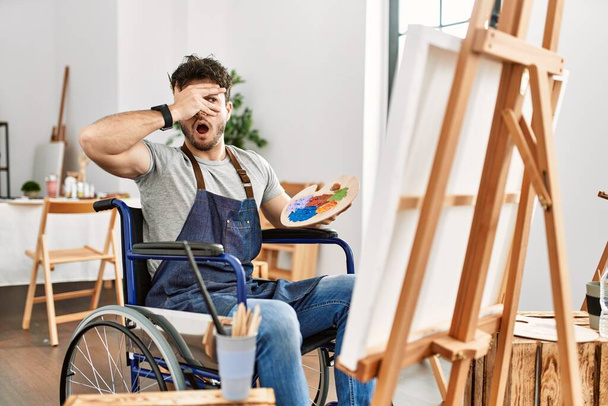 Junger hispanischer Mann, der im Rollstuhl im Kunstatelier sitzt und schockiert Gesicht und Augen mit der Hand bedeckt, blickt mit verlegenem Gesichtsausdruck durch Finger.  - Foto, Bild