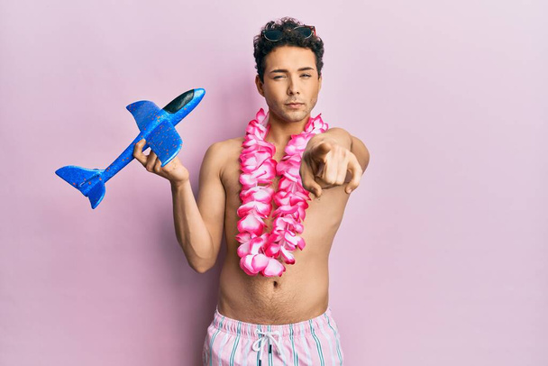 Young handsome man wearing swimsuit and hawaiian lei holding airplane toy pointing with finger to the camera and to you, confident gesture looking serious  - Fotografie, Obrázek