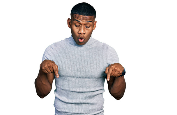 Young black man wearing casual t shirt pointing down with fingers showing advertisement, surprised face and open mouth  - 写真・画像