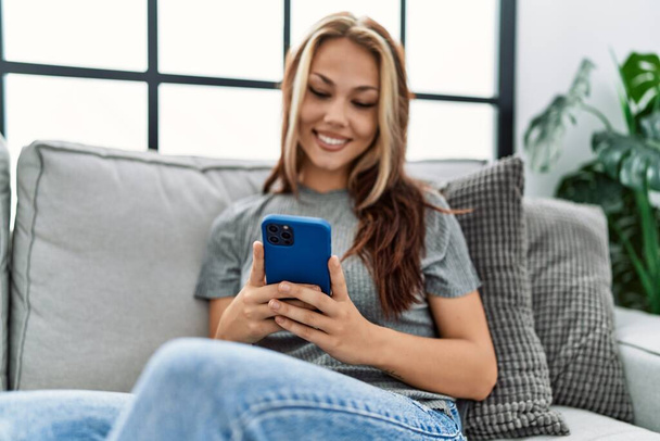 Jovem caucasiana usando smartphone sentado no sofá em casa. - Foto, Imagem