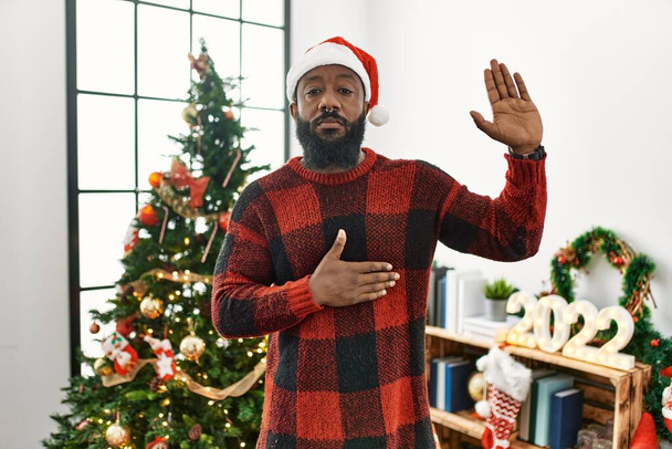 Αφροαμερικάνος που φοράει καπέλο Άη Βασίλη στέκεται δίπλα στο χριστουγεννιάτικο δέντρο βρίζοντας με το χέρι στο στήθος και την ανοιχτή παλάμη, δίνοντας όρκο πίστης.  - Φωτογραφία, εικόνα