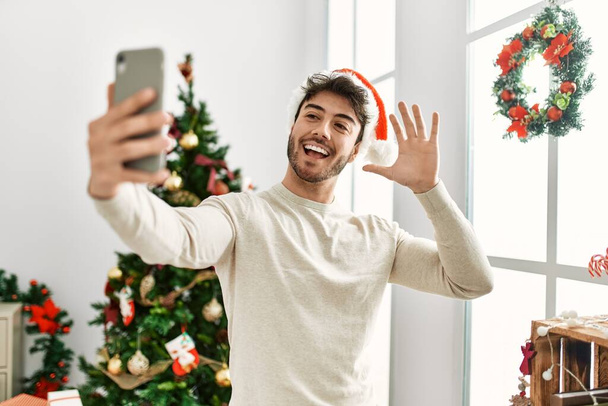 Young hispanic man wearing christmas hat making selfie by the smartphone at home. - Фото, изображение