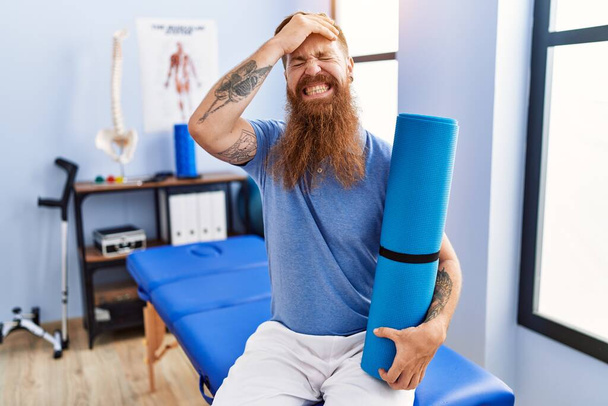 Redhead man with long beard holding yoga mat at rehabilitation clinic stressed and frustrated with hand on head, surprised and angry face  - Valokuva, kuva