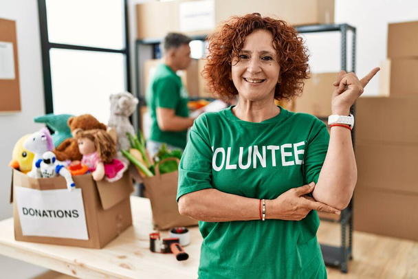 Middle age woman wearing volunteer t shirt at donations stand with a big smile on face, pointing with hand and finger to the side looking at the camera.  - 写真・画像