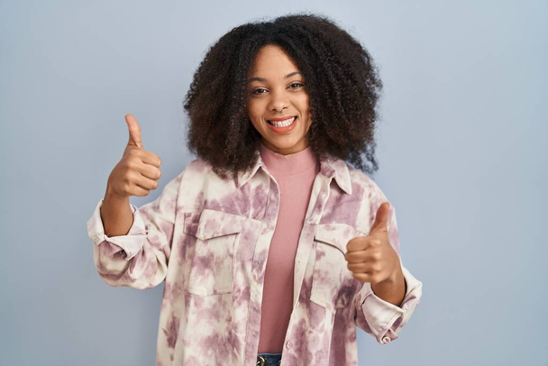 青い背景に立つ若いアフリカ系アメリカ人女性は、手で積極的なジェスチャーを行い、笑顔と幸せを親指アップ。陽気な表情と勝者のジェスチャー.  - 写真・画像