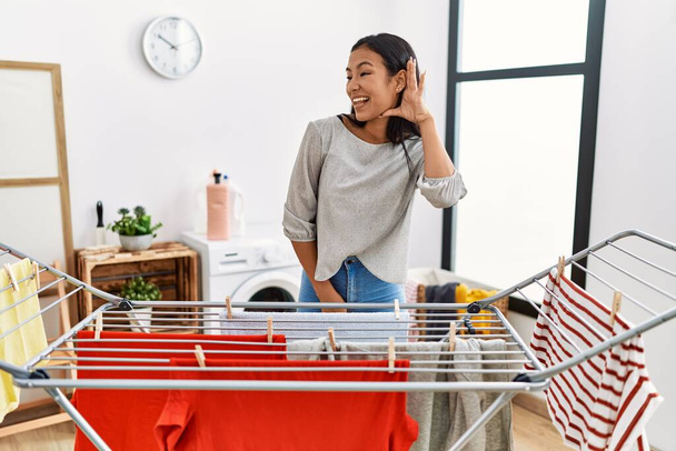 Young hispanic woman putting fresh laundry on clothesline smiling with hand over ear listening an hearing to rumor or gossip. deafness concept.  - Photo, Image