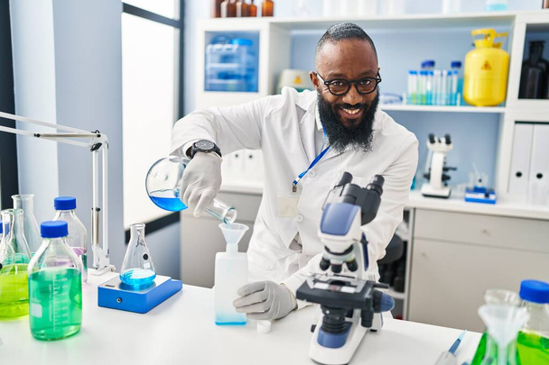 Jonge Afro-Amerikaanse man draagt wetenschapper uniform meten vloeistof in laboratorium - Foto, afbeelding