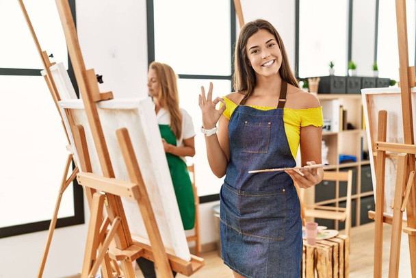 Young hispanic woman at art classroom doing ok sign with fingers, smiling friendly gesturing excellent symbol  - Foto, Imagem