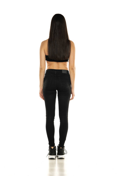 Rear view of a young woman in black jeans and short top posing on a white background - Foto, Imagen