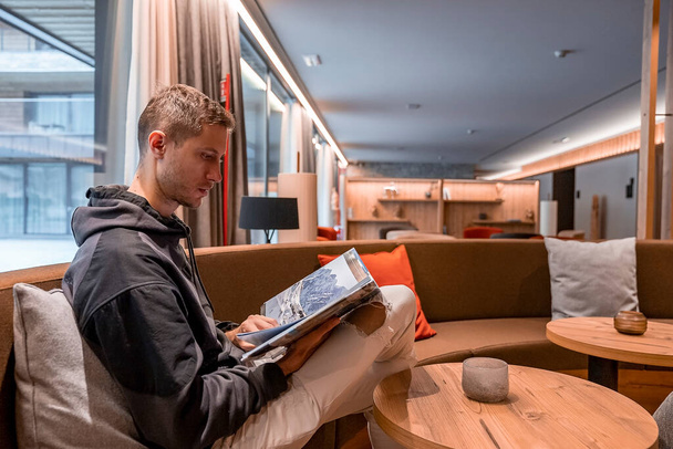Man reading travel magazine while sitting in lobby. Young male is looking through catalog while relaxing on sofa. He is resting in luxurious lounge at resort. - Fotografie, Obrázek