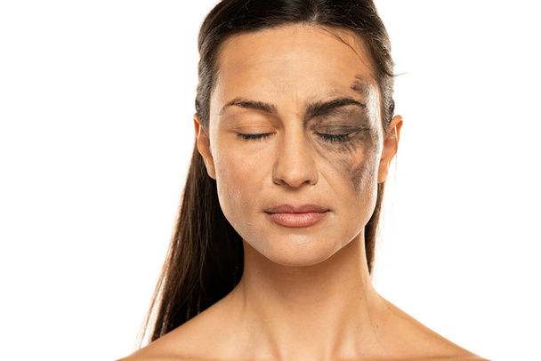 eine junge Frau mit geschlossenen Augen posiert mit halb verschmutztem und halb geschminktem Gesicht auf weißem Hintergrund. - Foto, Bild