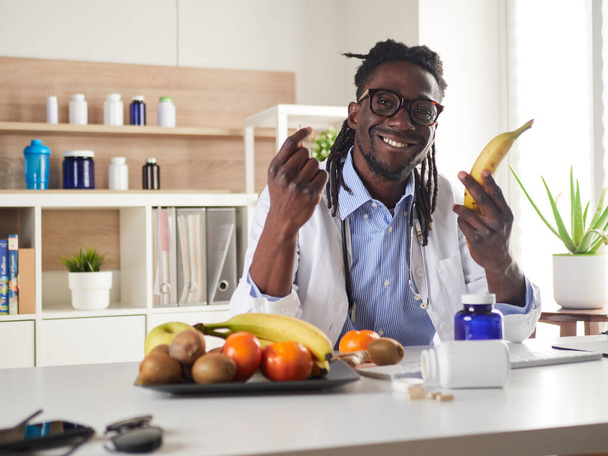 Afroamerikan beslenme uzmanı kameraya bakıyor ve konsültasyonda sağlıklı meyveler gösteriyor. - Fotoğraf, Görsel
