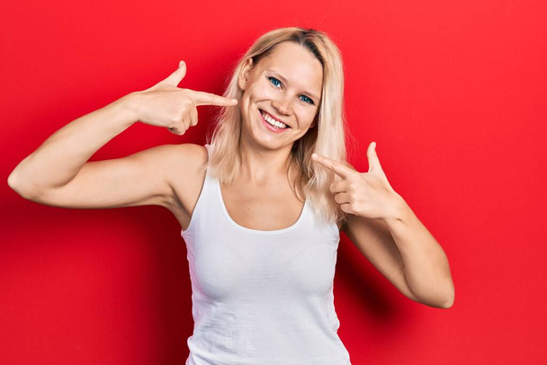 Beautiful caucasian blonde woman wearing casual white t shirt smiling cheerful showing and pointing with fingers teeth and mouth. dental health concept.  - Foto, Imagen