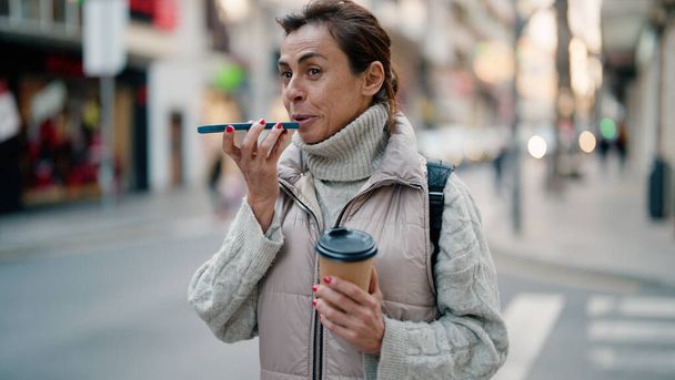 Hispanische Frau mittleren Alters spricht auf dem Smartphone und trinkt Kaffee auf der Straße - Foto, Bild
