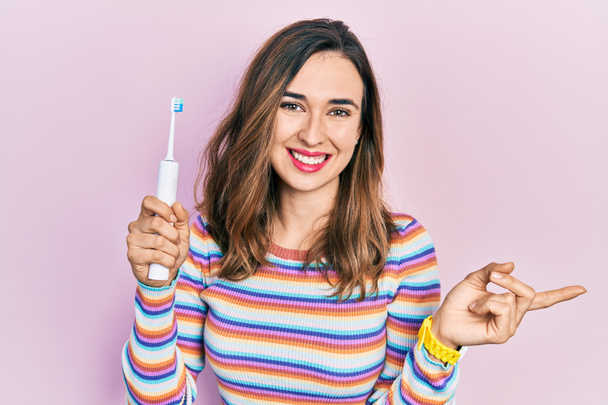 Giovane ragazza ispanica che tiene lo spazzolino da denti elettrico sorridente felice indicando con mano e dito di lato  - Foto, immagini