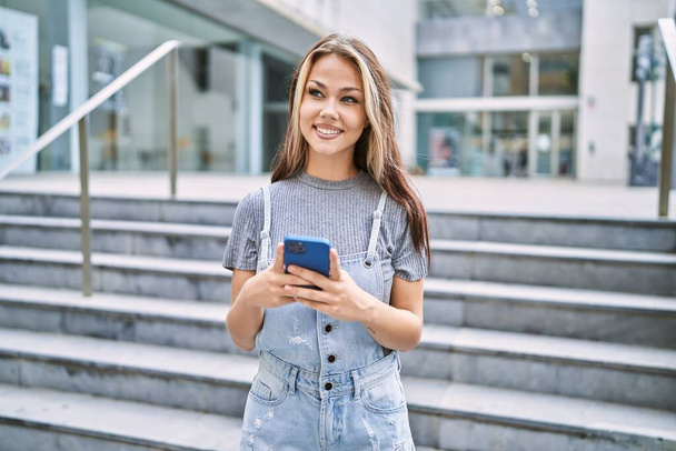 Νεαρή καυκάσια κοπέλα χαμογελώντας ευτυχισμένη χρησιμοποιώντας το smartphone στην πόλη. - Φωτογραφία, εικόνα