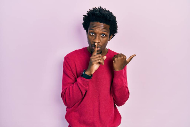 Jonge Afro-Amerikaanse man draagt casual kleding en vraagt om stil te zijn met de vinger op de lippen wijzend met de hand naar de zijkant. stilte en geheim concept.  - Foto, afbeelding