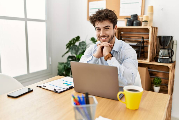 Νεαρός Ισπανός επιχειρηματίας χαμογελά ευτυχισμένος δουλεύοντας στο γραφείο. - Φωτογραφία, εικόνα