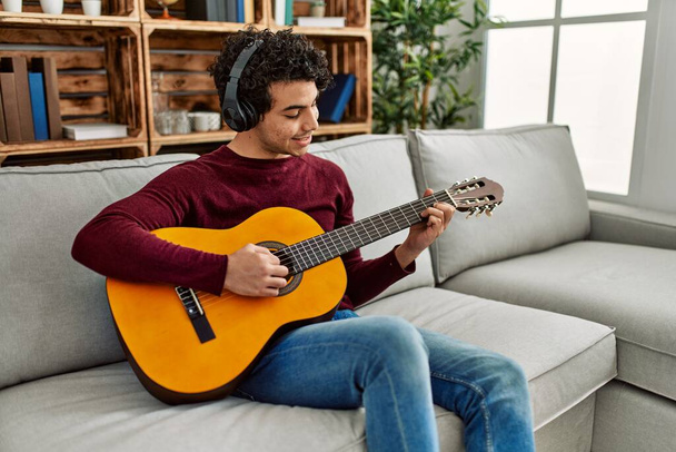 Jeune homme hispanique jouant de la guitare classique assis sur le canapé à la maison. - Photo, image