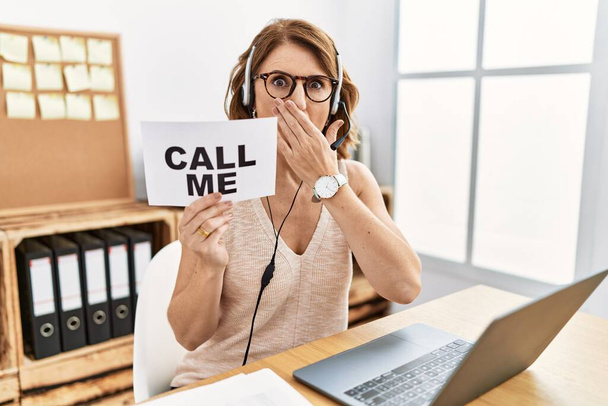 Middle age brunette woman wearing operator headset holding call me banner shocked covering mouth with hands for mistake. secret concept.  - Photo, image