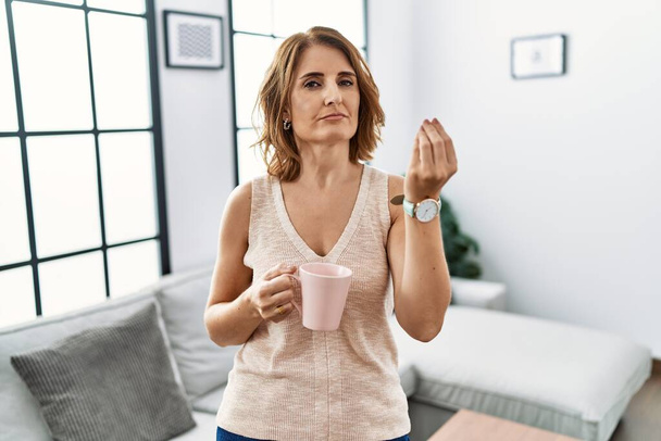 Frau mittleren Alters trinkt zu Hause eine Tasse Kaffee und macht italienische Geste mit selbstbewusstem Gesichtsausdruck  - Foto, Bild