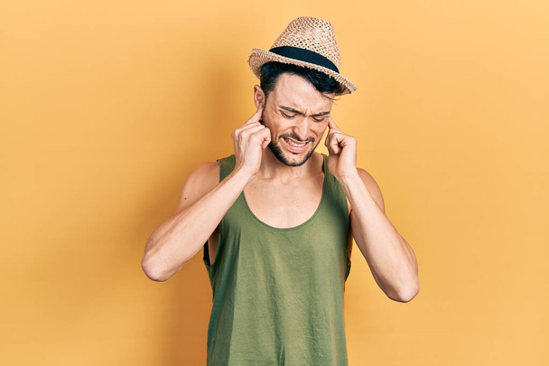 A fiatal spanyol férfi nyári kalapot visel, ujjakkal takarta be a fülét, bosszantó kifejezéssel a hangos zene zajára. süket fogalom.  - Fotó, kép