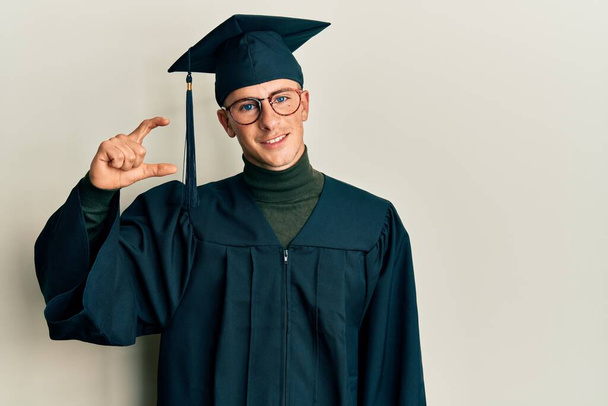 Young caucasian man wearing graduation cap and ceremony robe smiling and confident gesturing with hand doing small size sign with fingers looking and the camera. measure concept.  - Photo, Image