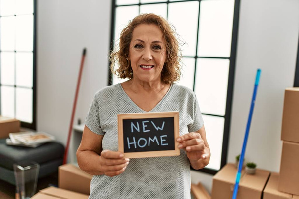 Mulher caucasiana de meia idade sorrindo feliz segurando novo quadro-negro casa em casa. - Foto, Imagem