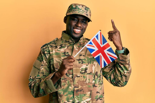 Young african american man wearing army uniform holding united kingdom flag smiling with an idea or question pointing finger with happy face, number one  - Photo, Image