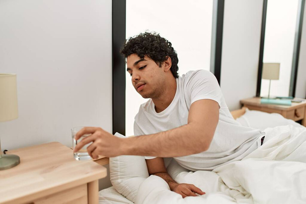 Jonge Spaanse man drinkwater zittend op het bed in de slaapkamer. - Foto, afbeelding