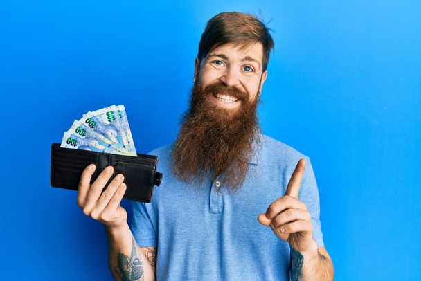 Redhead man with long beard holding wallet with south african rand banknotes smiling happy pointing with hand and finger to the side  - Φωτογραφία, εικόνα