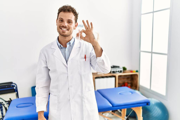 Guapo joven que trabaja en la clínica de recuperación del dolor sonriendo positiva haciendo signo de ok con la mano y los dedos. expresión exitosa.  - Foto, imagen