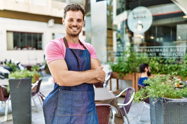Young hispanic waiter wearing apron standing with arms crossed gesture at coffee shop. - Photo, Image