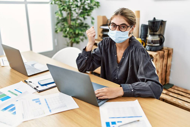 Young blonde woman working at the office wearing safety mask screaming proud, celebrating victory and success very excited with raised arm  - Foto, Imagem