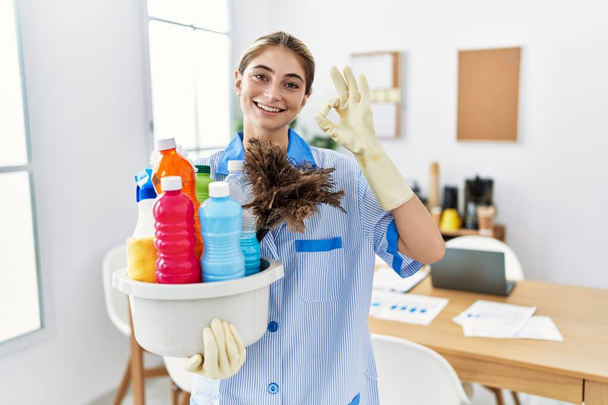 Young blonde woman wearing cleaner uniform holding cleaning products smiling positive doing ok sign with hand and fingers. successful expression.  - Photo, Image
