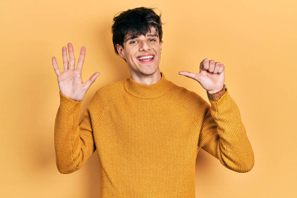Jóképű hipszter fiatalember alkalmi sárga pulóvert visel, mutatva és mutatva a hatos számú ujjaival, miközben magabiztosan és boldogan mosolyog..  - Fotó, kép