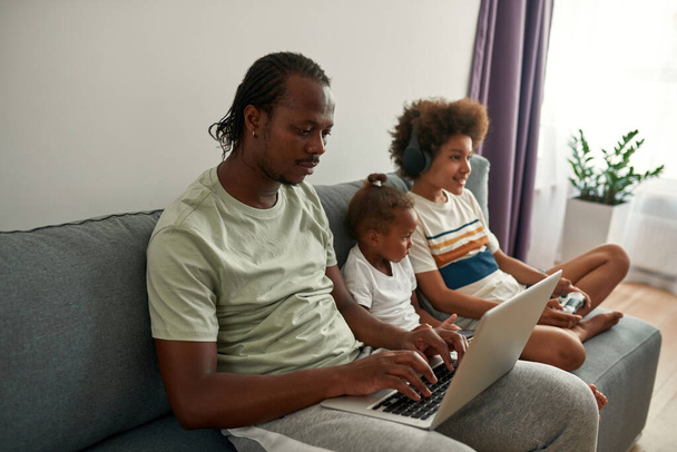 Black family using digital devices on sofa at home - Фото, изображение