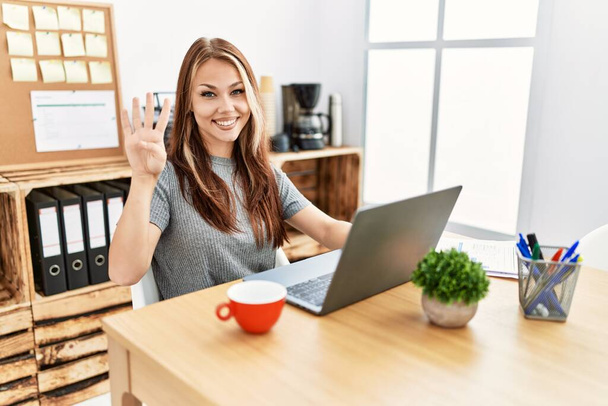 Молода брюнетка працює в офісі з ноутбуком, показуючи і вказуючи пальцями номер чотири, посміхаючись впевнено і щасливо
.  - Фото, зображення