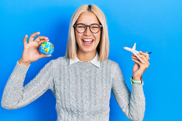 Beautiful blonde woman holding airplane toy and world ball smiling and laughing hard out loud because funny crazy joke.  - Foto, Imagen