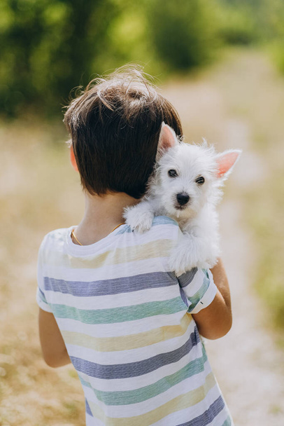 Friendship of a child with a dog. Happy boy holding a West Highland White Terrier puppy in his arms. The child walks with his puppy in the summer in the park. The boy's favorite puppy in nature. - Foto, Imagem