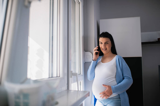 Une femme caucasienne enceinte utilise un téléphone portable, parle, sourit et regarde la caméra pendant qu'elle se tient à côté de la porte du salon. Sa main gauche est sur son ventre et elle tient son téléphone portable avec sa droite. - Photo, image