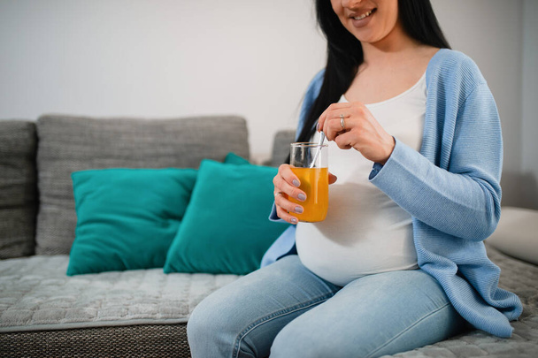 Черноволосая беременная кавказка смотрит на стакан апельсинового сока и смешивает его с ложкой. Улыбающаяся женщина сидит в гостиной на диване. На ней белая футболка и голубой свитер..  - Фото, изображение