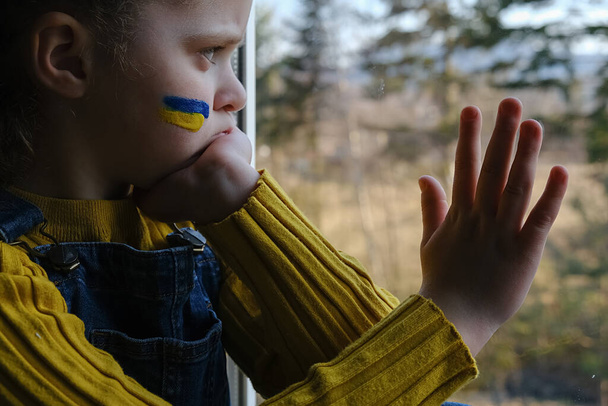 Side view of upset sad Ukrainian little girl child sitting alone at window. State support in war. United Europe against Russia. Hope for the world. Anxiety and anticipation. Save Ukraine - Foto, imagen